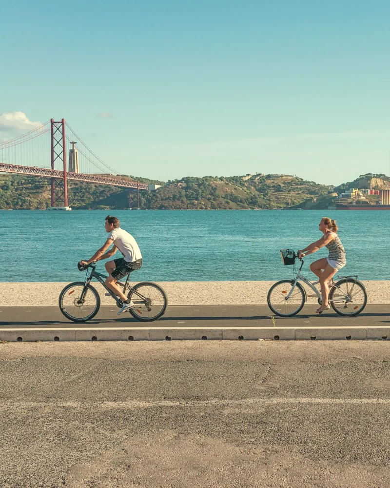 People cycling along the Tagus riverside in Lisbon, with the 25 de Abril Bridge behind