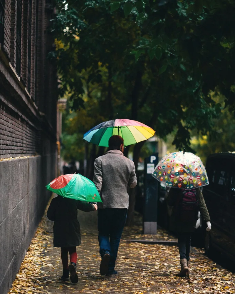 An adult and two children with colourful umbrellas walking down a leafy Madrid street in autumn
